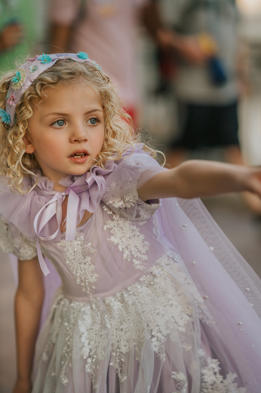 A young girl wears a soft tulle lavender cape tied at the shoulders with a satin ribbon. The cape has beautiful pearls placed all over and a ruffle collar.