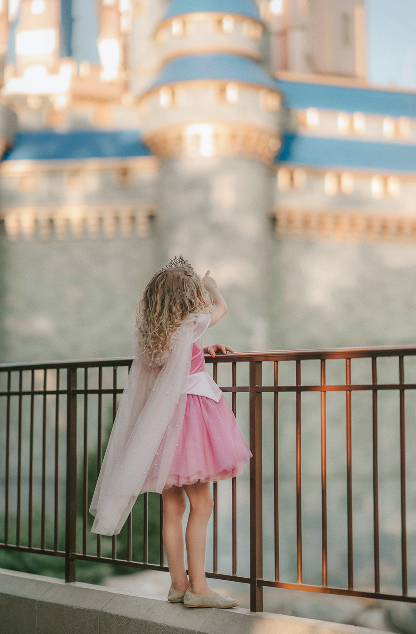 A young girl wears a soft tulle light pink cape tied at the shoulders with a satin ribbon. The cape has beautiful pearls placed all over and a ruffle collar.