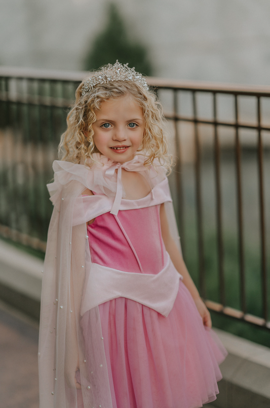 A young girl wears a soft tulle light pink cape tied at the shoulders with a satin ribbon. The cape has beautiful pearls placed all over and a ruffle collar.