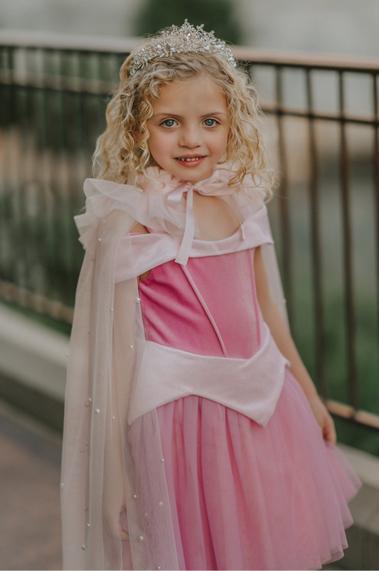 A young girl wears a soft tulle light pink cape tied at the shoulders with a satin ribbon. The cape has beautiful pearls placed all over and a ruffle collar.