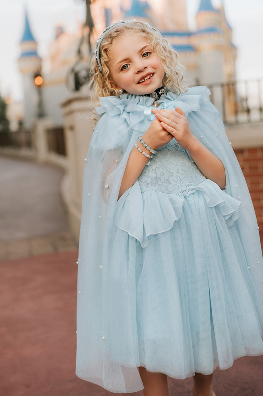A young girl wears a soft tulle light blue cape tied at the shoulders with a satin ribbon. The cape has beautiful pearls placed all over and a ruffle collar.