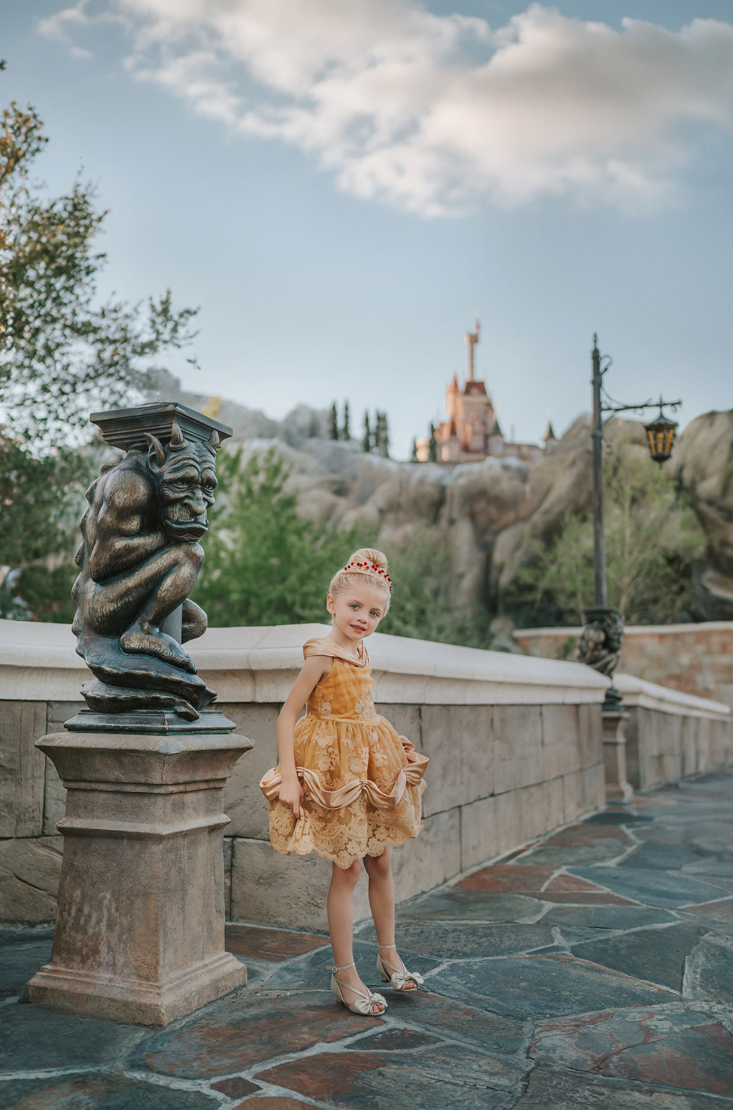A young girl with blonde hair stands outdoors, wearing the Portrait Petite Red Rose Princess twirl length gown, an intricately designed gold princess dress adorned with floral lace patterns. The satin off shoulder neckline is adorned with a gold jewel.