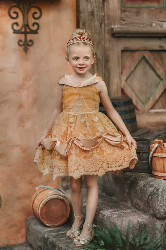 A young girl with blonde hair stands outdoors, wearing the Portrait Petite Red Rose Princess twirl length gown, an intricately designed gold princess dress adorned with floral lace patterns. The satin off shoulder neckline is adorned with a gold jewel.