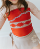 A young girl is wearing a bright red swim top with flower and pattern band. The tan-grey swimsuit bottom is adorned with a tan hibiscus flower—it's the Polynesian Princess Rashguard Swim Set made from SPF 50 material.
