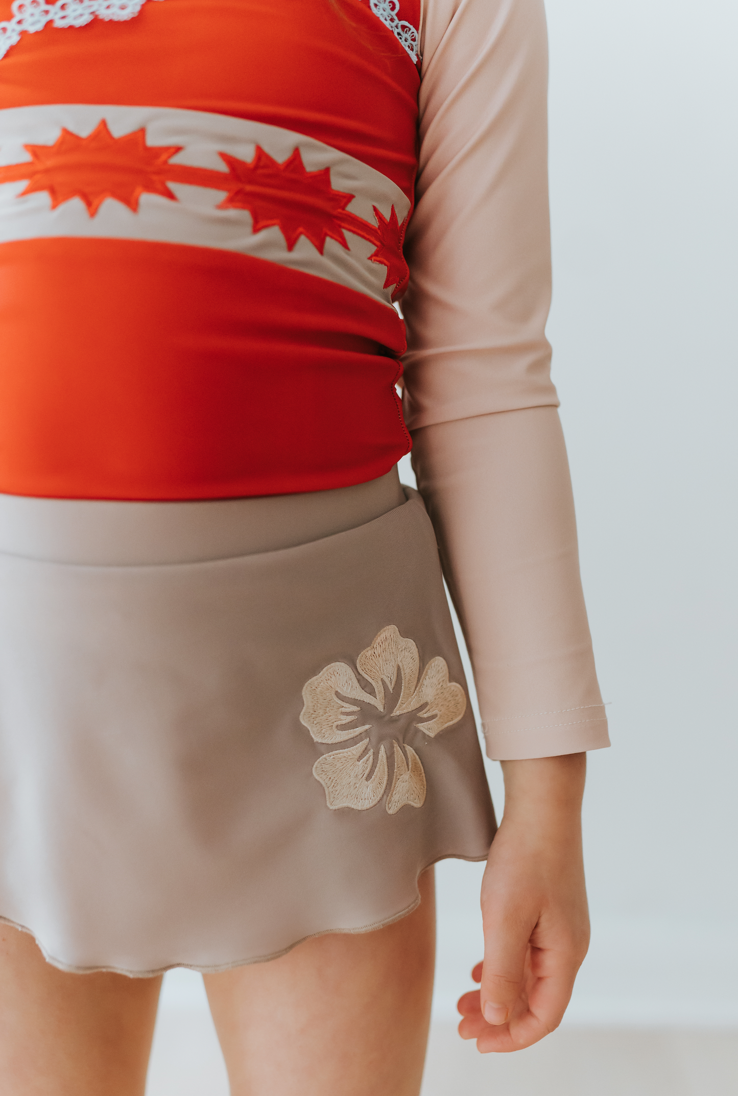 A young girl is wearing a bright red swim top with flower and pattern band. The tan-grey swimsuit bottom is adorned with a tan hibiscus flower—it's the Polynesian Princess Rashguard Swim Set made from SPF 50 material.