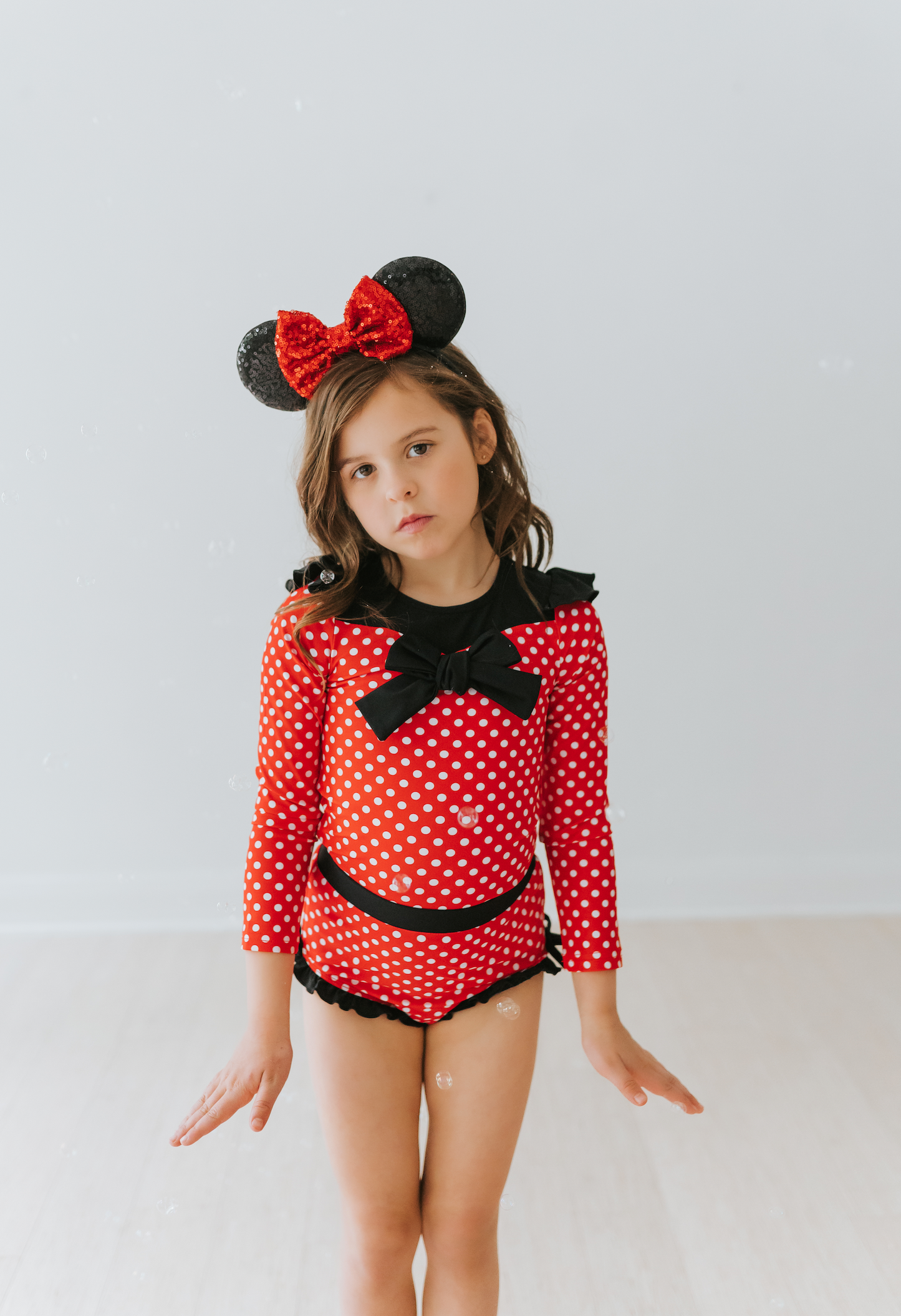 A young girl is wearing our Miss Mouse Rashguard Swim set. This 2pc swim has SPF 50. The top is red with white polka dots. There is a large black bow in the center and small ruffles on the shoulder. The bottoms have black ruffles along the edge.