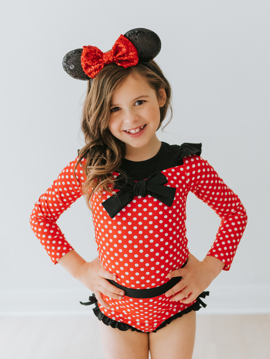 A young girl is wearing our Miss Mouse Rashguard Swim set. This 2pc swim has SPF 50. The top is red with white polka dots. There is a large black bow in the center and small ruffles on the shoulder. The bottoms have black ruffles along the edge.