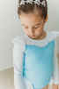 A young girl is wearing a long sleeve light blue and white rash guard swim set. The top is white ruffles at the shoulder and light blue floral ribbon trim across the chest. The modest bottoms are light blue with a white partial skirt that wraps around the back and is open in the front.