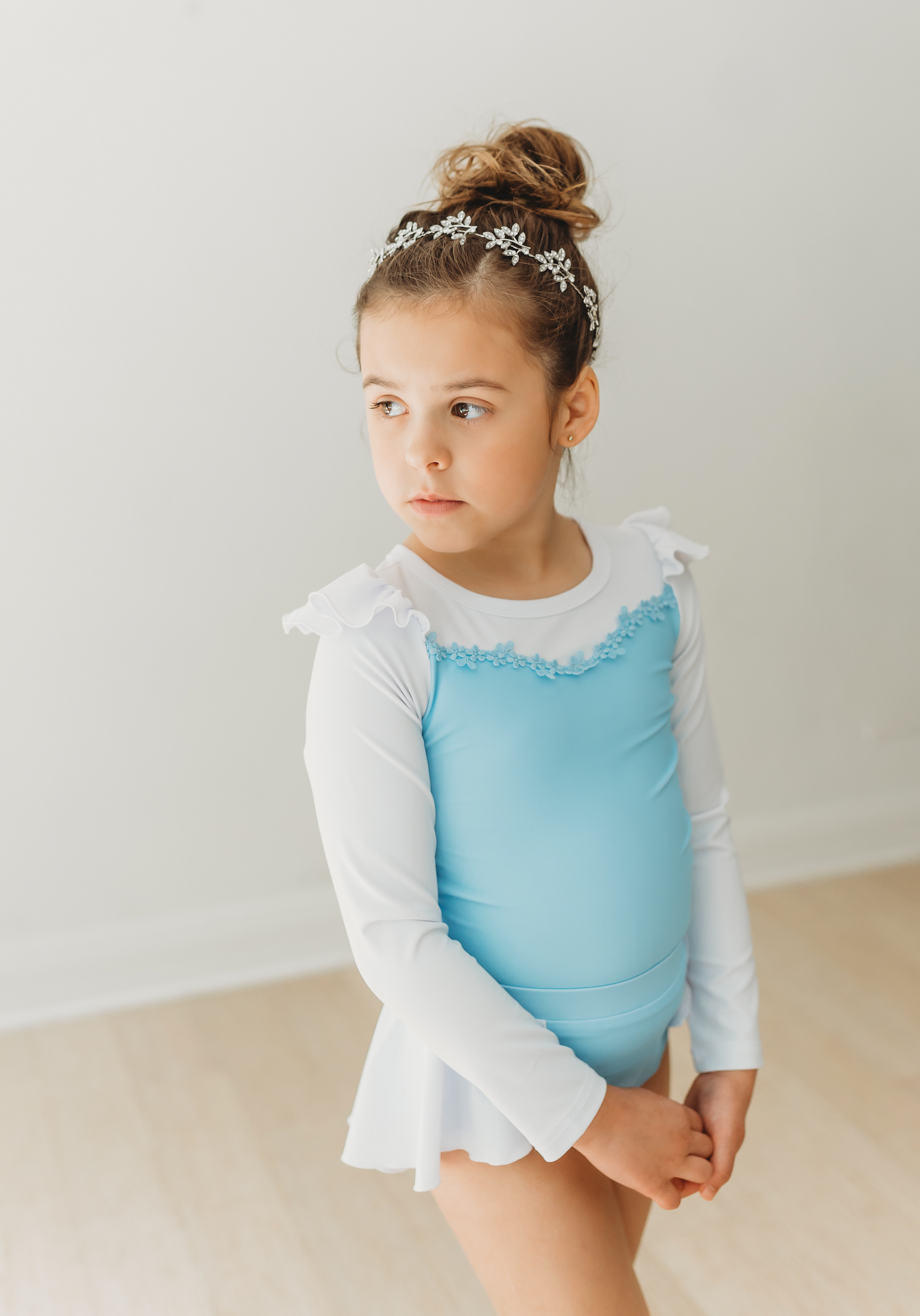 A young girl is wearing a long sleeve light blue and white rash guard swim set. The top is white ruffles at the shoulder and light blue floral ribbon trim across the chest. The modest bottoms are light blue with a white partial skirt that wraps around the back and is open in the front.