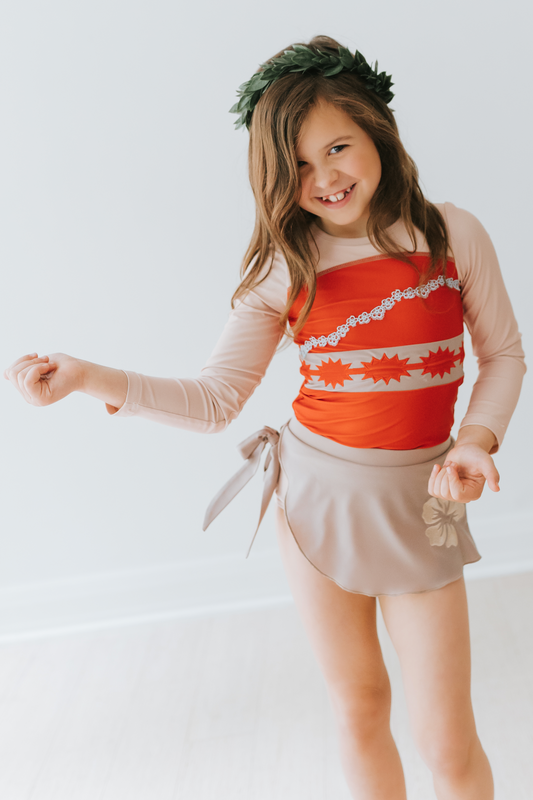 A young girl is wearing a bright red swim top with flower and pattern band. The tan-grey swimsuit bottom is adorned with a tan hibiscus flower—it's the Polynesian Princess Rashguard Swim Set made from SPF 50 material.
