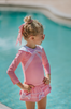 A young girl is wearing a long sleeve 2 piece pink rash guard swim set. There is white collar with a pink bow at the neck. Two lighter pink stripes are on the chest like ribbon tails. The bottom is a skirt with bows light pink bows at the hem.