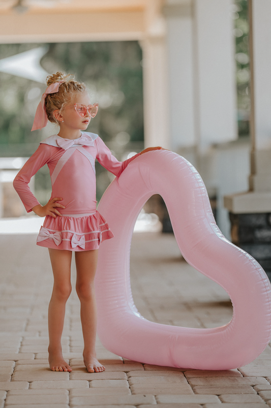 A young girl is wearing a long sleeve 2 piece pink rash guard swim set. There is white collar with a pink bow at the neck. Two lighter pink stripes are on the chest like ribbon tails. The bottom is a skirt with bows light pink bows at the hem.