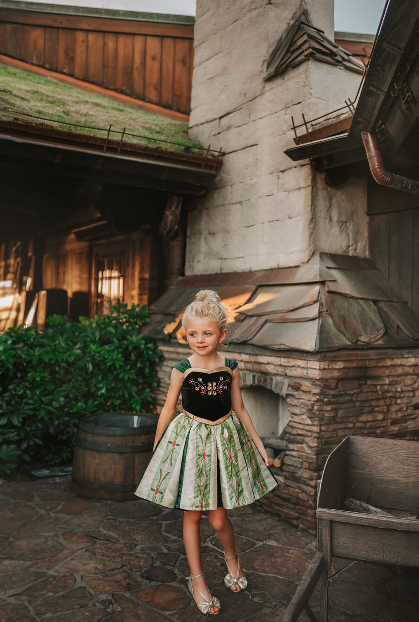 A young girl with blonde hair wearing the Portrait Petites Coronation Ice Princess Twirl Length Gown adorned with custom embroidery in green and black. The black stretch velvet bodice is adorned with golden jewels and flower embroidery. The skirt is shades of green with pink and green embroidered designs.