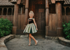 A young girl with blonde hair wearing the Portrait Petites Coronation Ice Princess Twirl Length Gown adorned with custom embroidery in green and black. The black stretch velvet bodice is adorned with golden jewels and flower embroidery. The skirt is shades of green with pink and green embroidered designs.