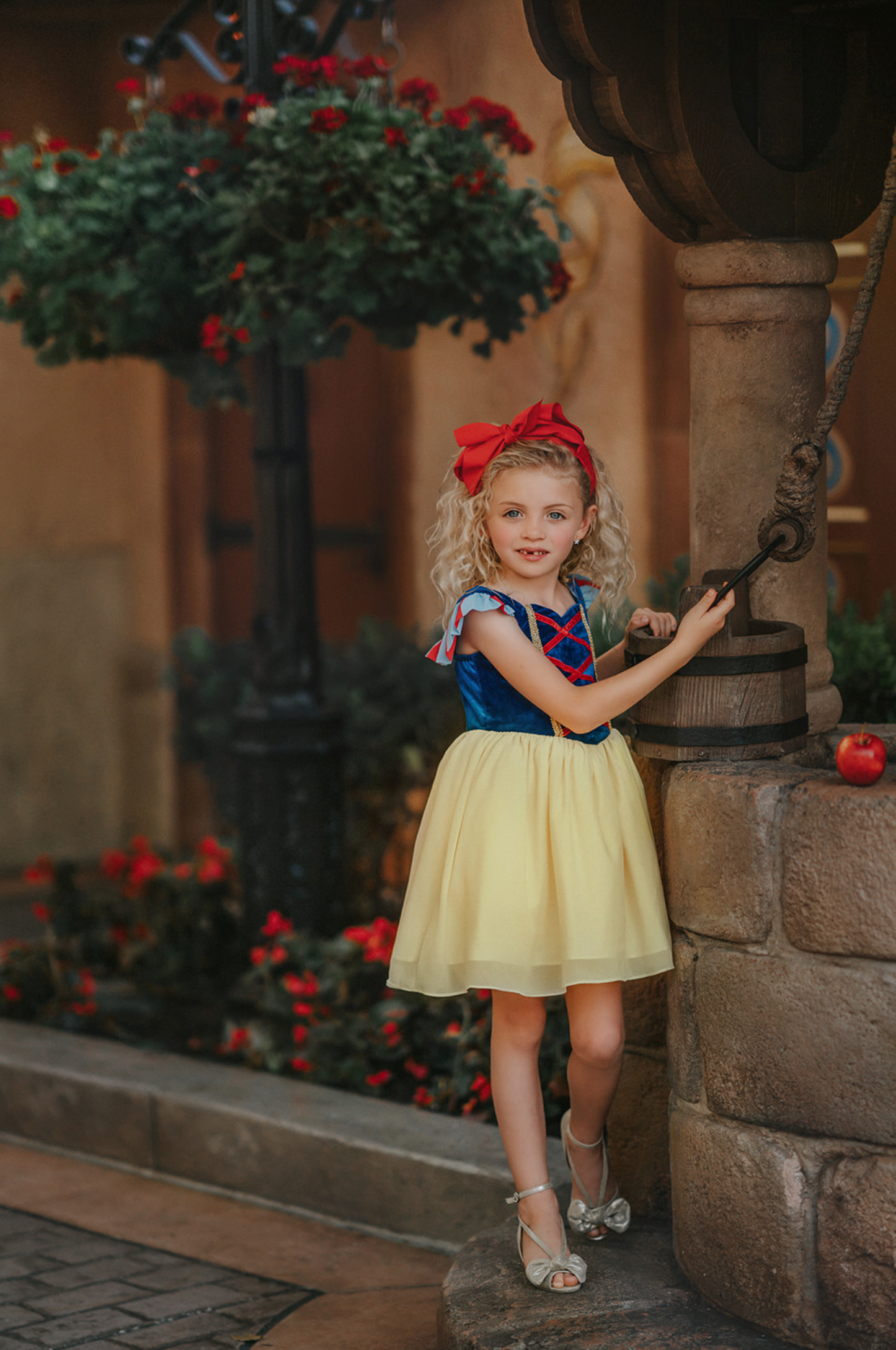 A young girl is wearing the Portrait Petite Twirl Length Snow White Gown. The gown is velvet blue and a light yellow skirt with a red ribbon criss cross accent on the bodice. The dress is sleeveless with a red and blue flutter ruffle on the top.