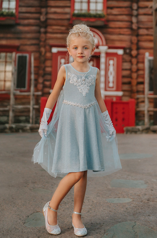 A young girl is dressed in the sparkly light blue Portrait Petite Ice Queen Gown in twirl length adorned with intricate custom lace at the neckline with rhinestones. A light blue cape with silver sparkle snowflakes is buttoned to the back of the gown.
