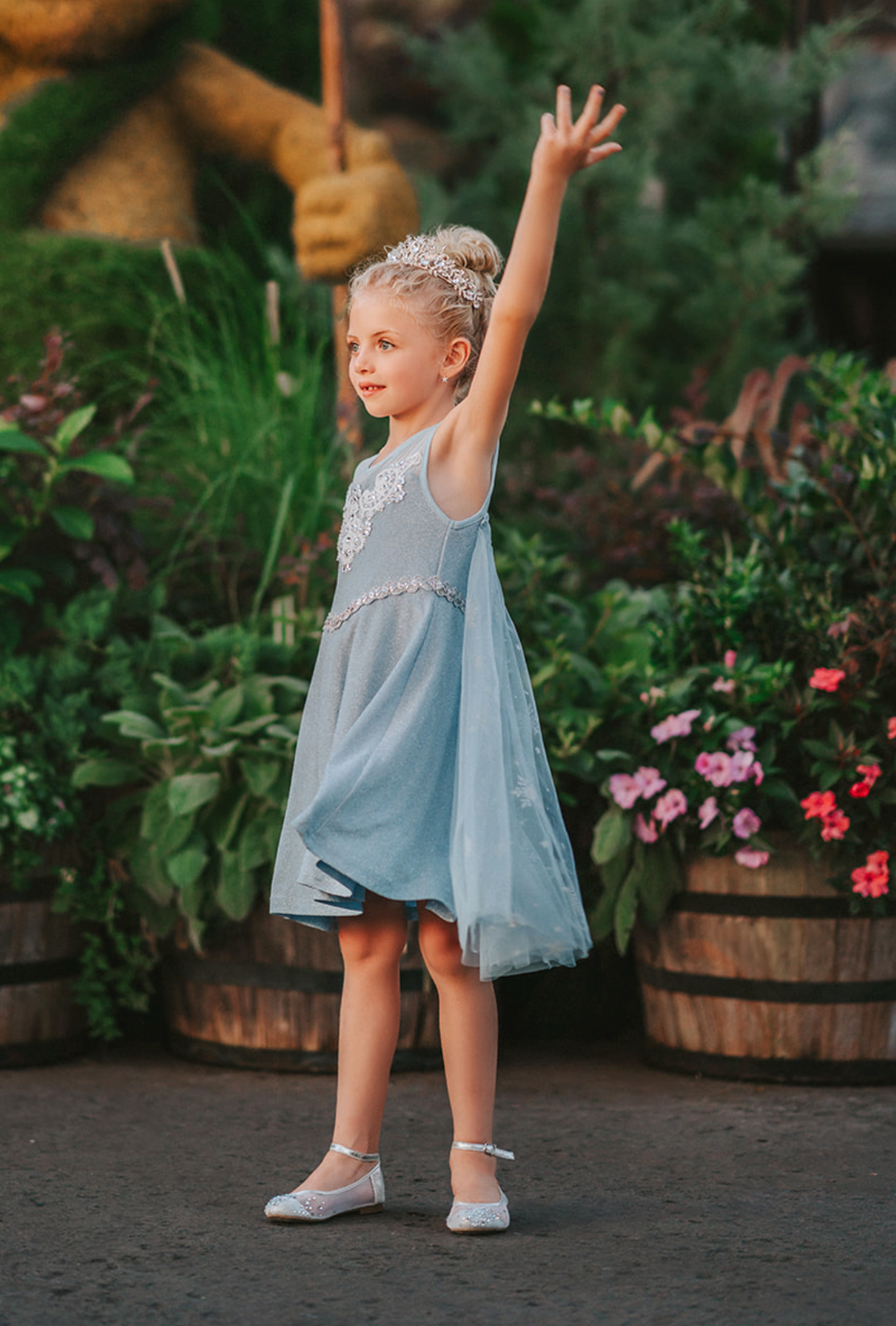 A young girl is dressed in the sparkly light blue Portrait Petite Ice Queen Gown in twirl length adorned with intricate custom lace at the neckline with rhinestones. A light blue cape with silver sparkle snowflakes is buttoned to the back of the gown.