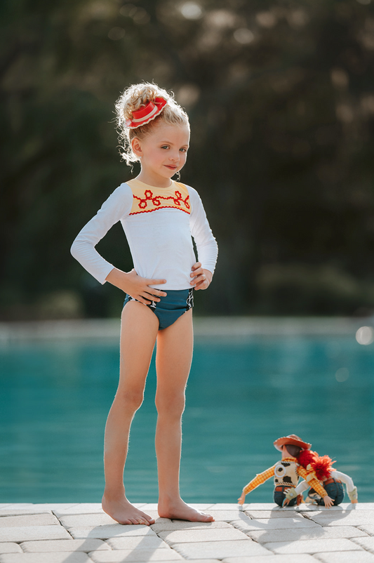 A young girl is wearing a 2 piece long sleeve rash guard swim set. The top is white with yellow on the upper chest. Across the yellow is red scrolling rick rack. The bottoms are a faux denim with black and white cow print panels.