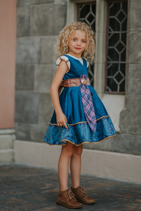 A young girl is wearing a long peacock blue green velvet twirl length dress. The velvet has silver sparkle. The sleeveless dress as off white accents on the shoulder. There is gold rope trim around the skirt. A Celtic pattern band in tan has a purple and blue plaid sash with a gold button accent. The base of the dress has a Celtic inspired pattern in shades of blue green.
