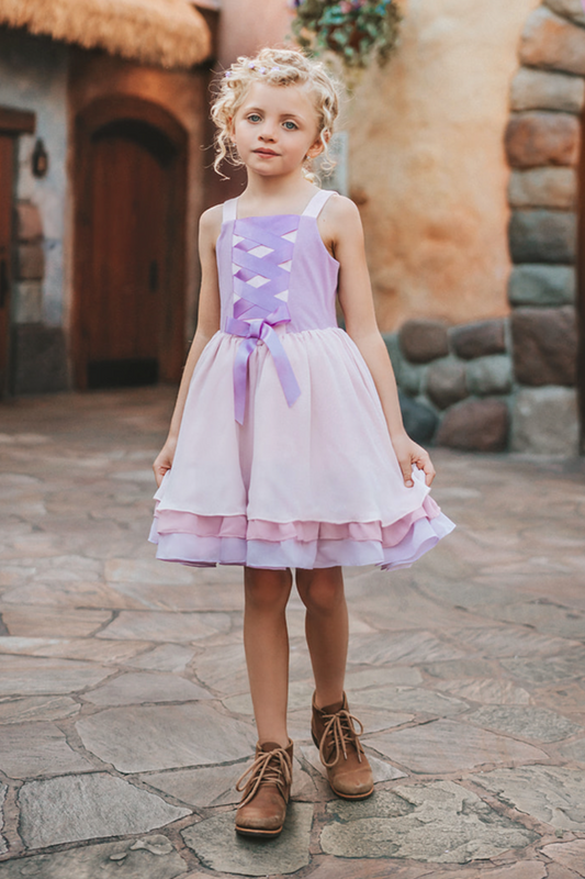 A young girl is adorned in the Portrait Petite Twirl Length Rapunzel Gown—a flowing, light violet princess dress featuring a purple ribbon corset design on the bodice. The skirts are 3 layers of light dusty purple, pink and lavender. The skirt is perfect for twirling and dancing in any enchanted land.