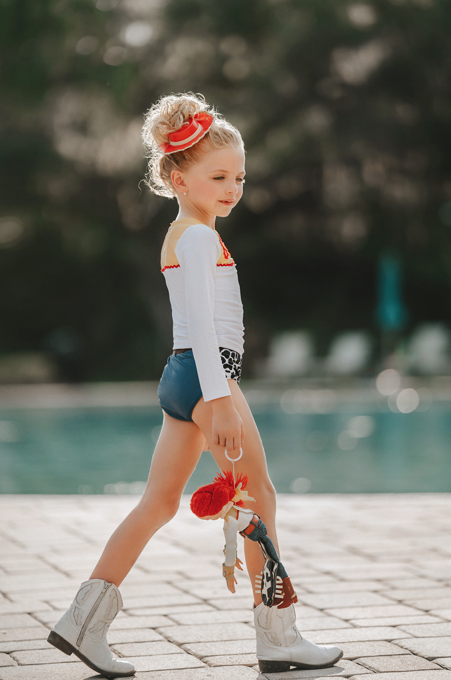 A young girl is wearing a 2 piece long sleeve rash guard swim set. The top is white with yellow on the upper chest. Across the yellow is red scrolling rick rack. The bottoms are a faux denim with black and white cow print panels.