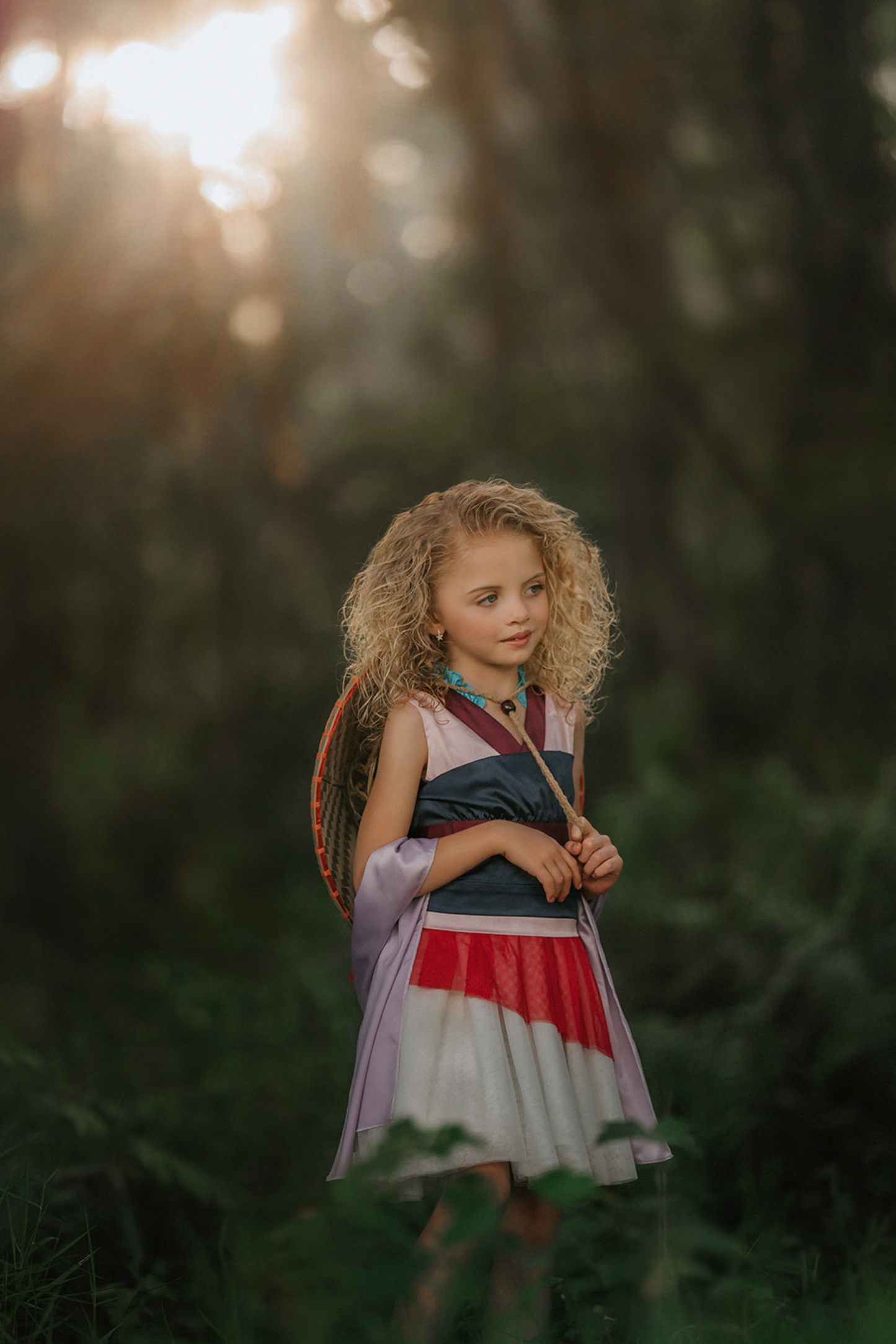 A young girl is wearing our Warrior Girl Twirl Length Portrait Petite Gown. The soft satin top is light pink with a maroon collar. The tulle skirt is white with an asymmetrical red layer. There is a navy blue corset piece that is removable. This dress also comes with a light purple scarf.