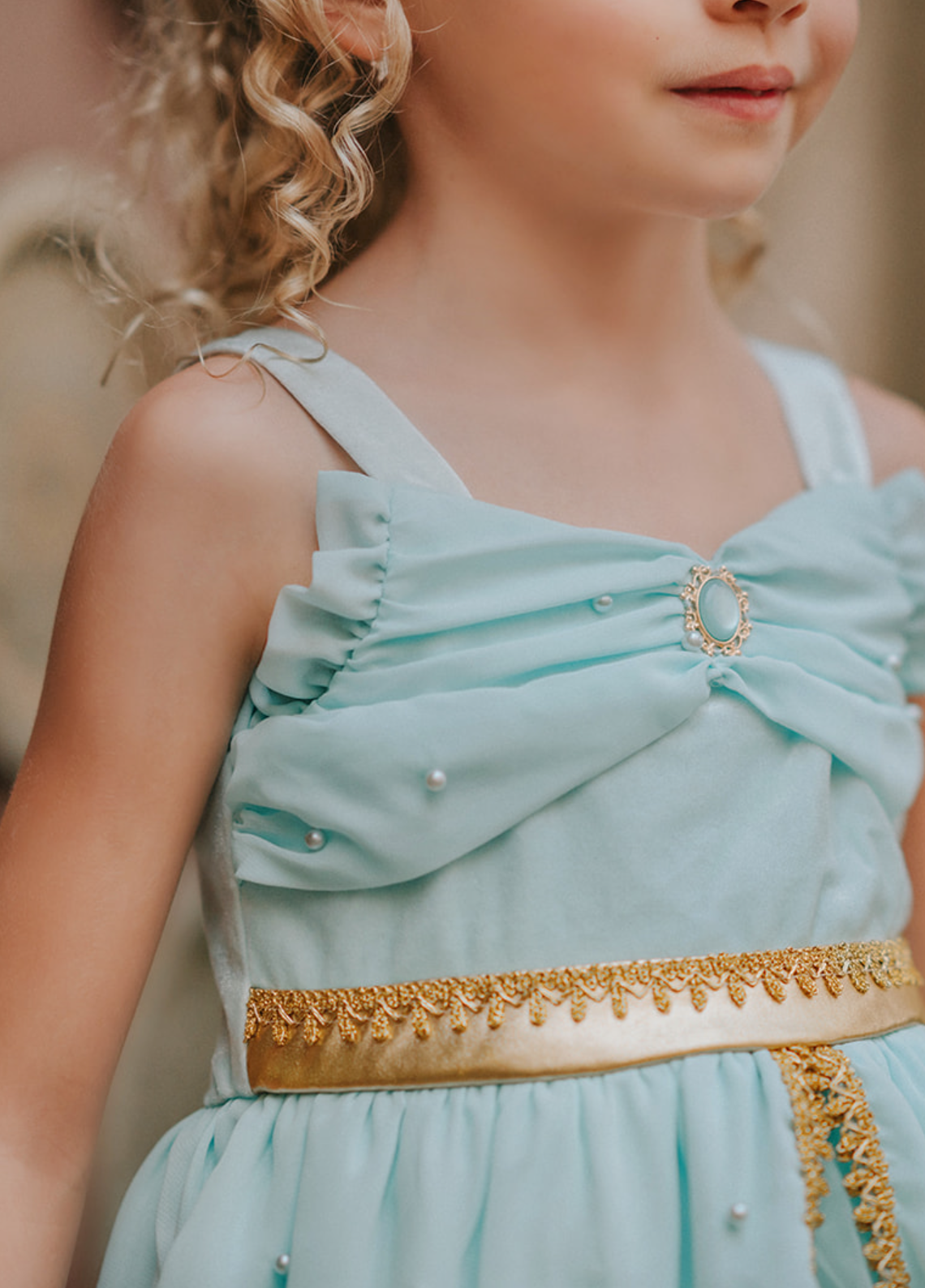 A young girl wears a light mint colored twirl length gown. The chiffon bodice has pearls adorned on it with a gold lace trim and waist. The layers of chiffon and tulle on the skirt have gold lace trim and pearls. This dress is stunning and will make any little one feel like an Arabian Princess.