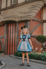 A young girl with curly blonde hair, wearing the Alice Tea Party Dress Reimagined in blue and white with a large black bow, stands in front of a quaint, thatched-roof cottage with red brick and wooden beams. She has a black headband with a bow on her head and is gazing thoughtfully into the distance.