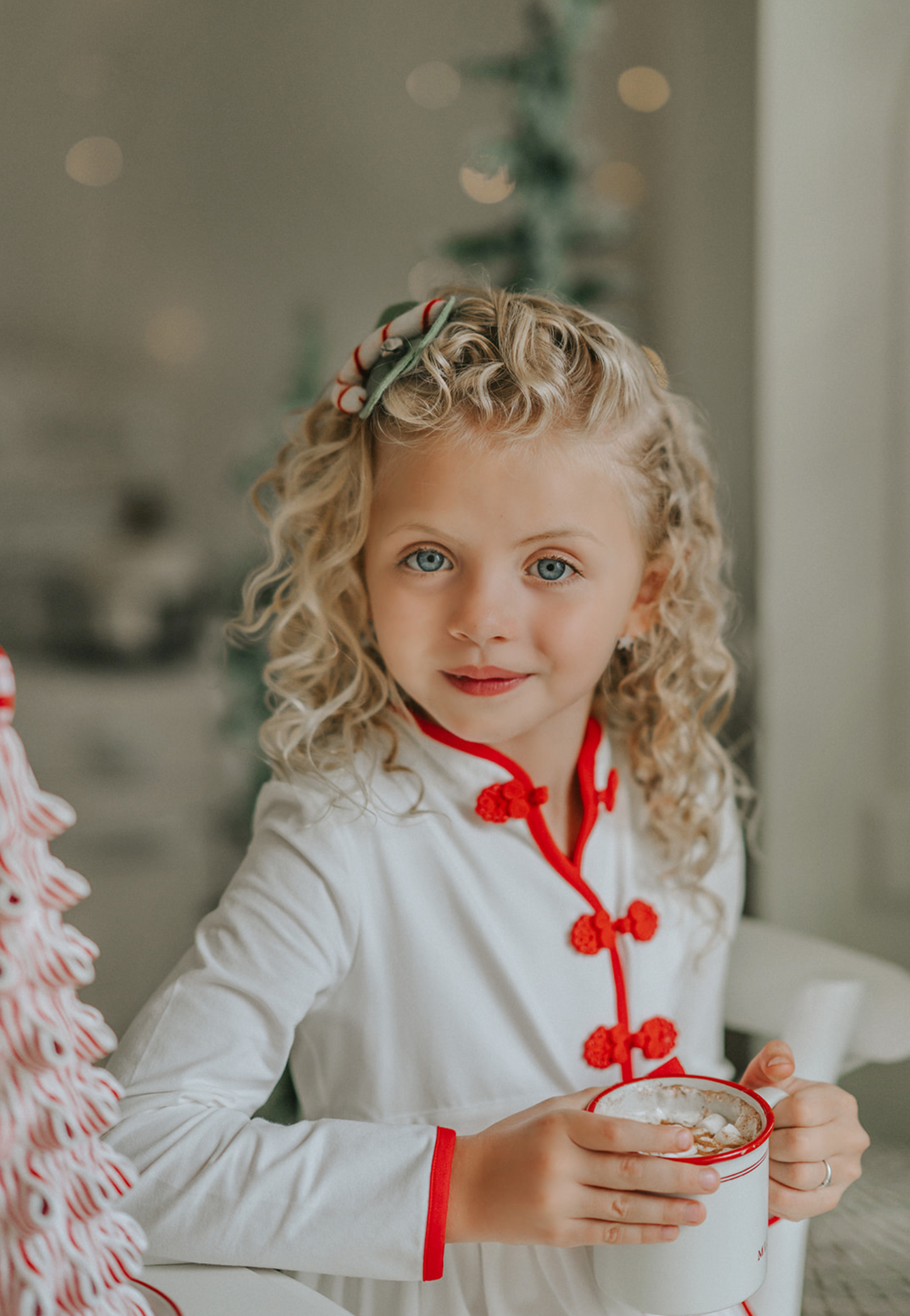 A young girl is wearing a white robe kimono. The buttery soft fabric is long sleeved with red piping trim. There are toggle buttons and a red ribbon tie at the waist. A perfect lounge piece to watch a White Christmas.