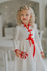 A young girl is wearing a white robe kimono. The buttery soft fabric is long sleeved with red piping trim. There are toggle buttons and a red ribbon tie at the waist. A perfect lounge piece to watch a White Christmas.