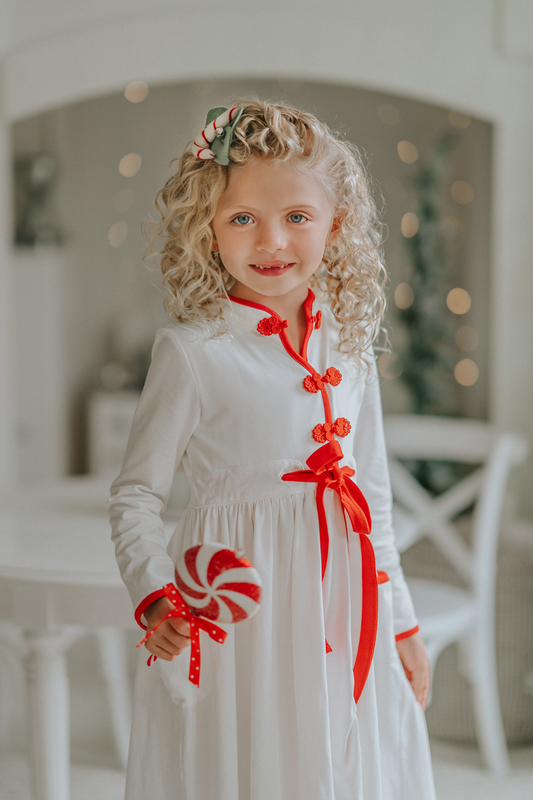 A young girl is wearing a white robe kimono. The buttery soft fabric is long sleeved with red piping trim. There are toggle buttons and a red ribbon tie at the waist. A perfect lounge piece to watch a White Christmas.