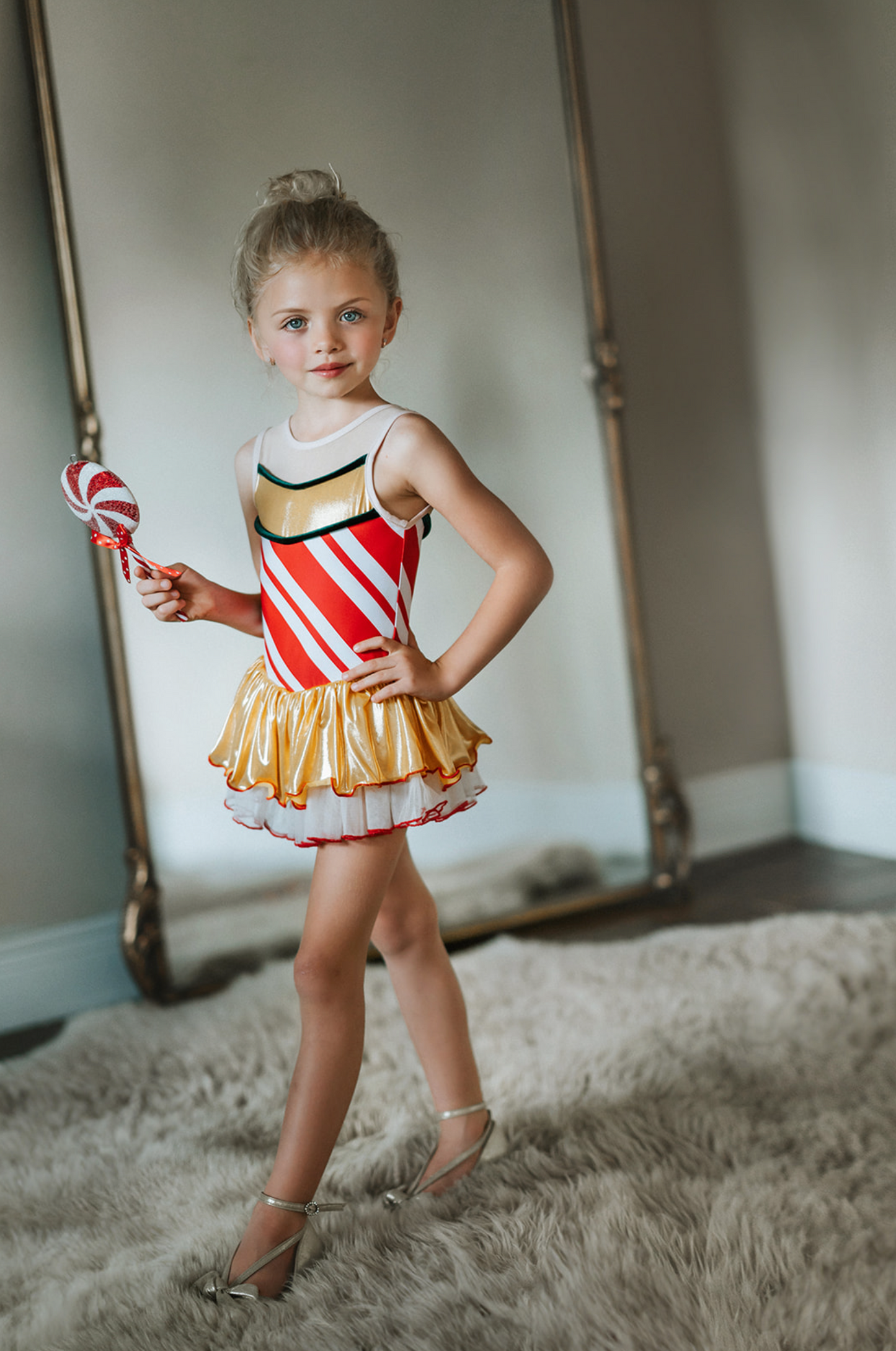 A young girl is wearing a skirted leotard. Red and white candy cane graphic fabric is paired with metallic gold and green velvet accents. The skirt has a metallic gold layer and a white layer below trimmed with red.