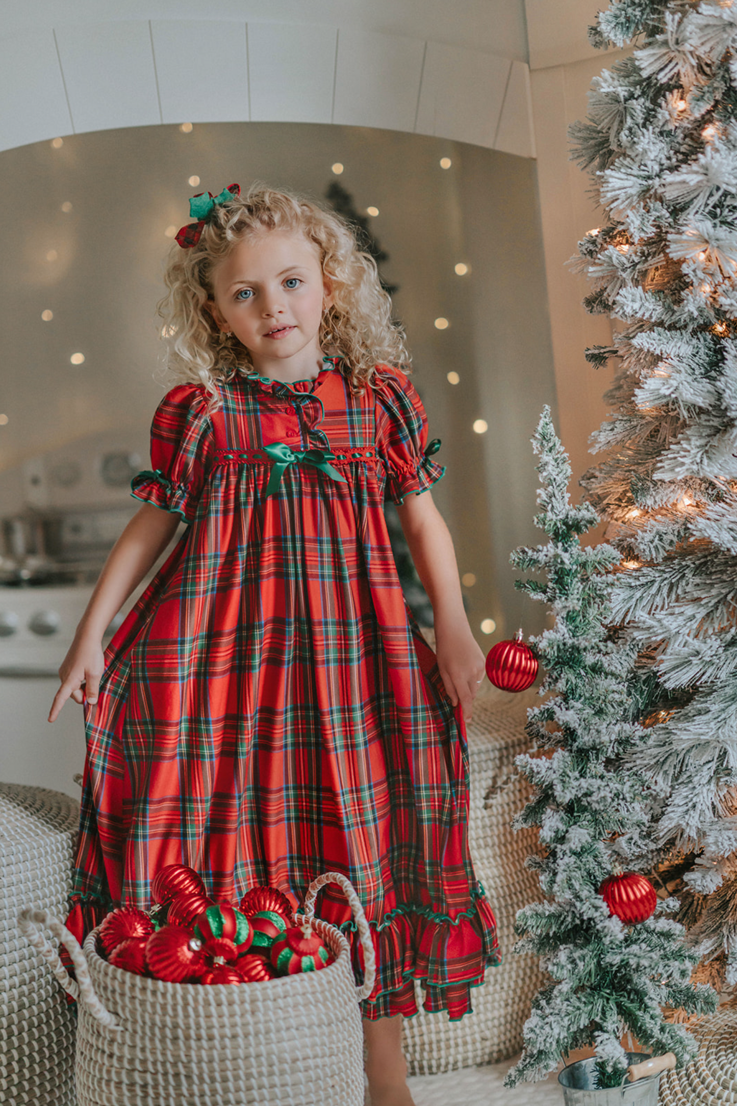 A young girl is wearing The Original Clara Nutcracker Gown in Red Plaid, featuring red plaid ruffles and a green bow. The plaid has white, green and black with red background. The buttery soft fabric is cozy and sensory friendly. The puff sleeves with ribbon detail and the ruffles at the hem add a traditional classic look.