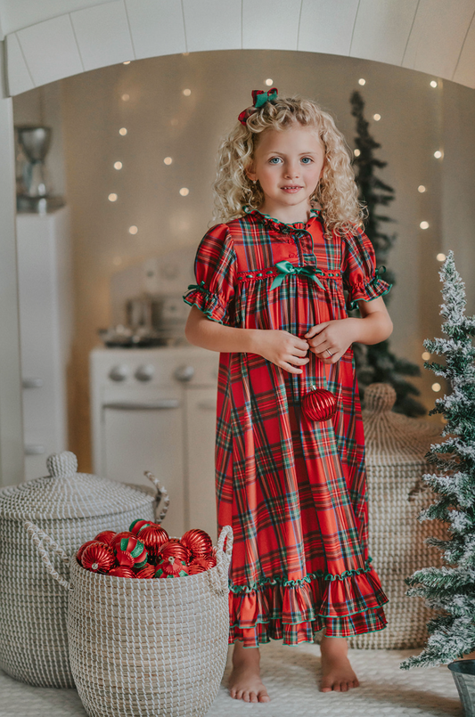 A young girl is wearing The Original Clara Nutcracker Gown in Red Plaid, featuring red plaid ruffles and a green bow. The plaid has white, green and black with red background. The buttery soft fabric is cozy and sensory friendly. The puff sleeves with ribbon detail and the ruffles at the hem add a traditional classic look.