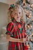 A young girl is wearing The Original Clara Nutcracker Gown in Red Plaid, featuring red plaid ruffles and a green bow. The plaid has white, green and black with red background. The buttery soft fabric is cozy and sensory friendly. The puff sleeves with ribbon detail and the ruffles at the hem add a traditional classic look.