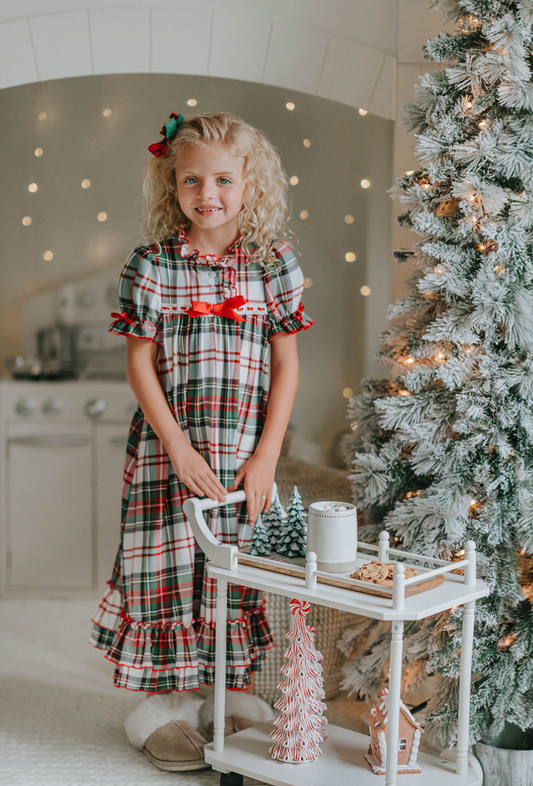 A young girl is wearing The Original Clara Nutcracker Gown in White Plaid, featuring white plaid ruffles and a bright red bow. The buttery soft fabric is cozy and sensory friendly. The puff sleeves with ribbon detail and the ruffles at the hem add a traditional classic look.