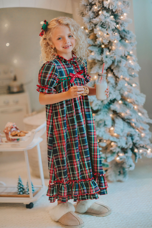 A young girl is wearing The Original Clara Nutcracker Gown in black plaid featuring black plaid ruffles and a red bow. The plaid has white, red and green with a black background. The buttery soft fabric is cozy and sensory friendly. The puff sleeves with ribbon detail and the ruffles at the hem add a traditional classic look.