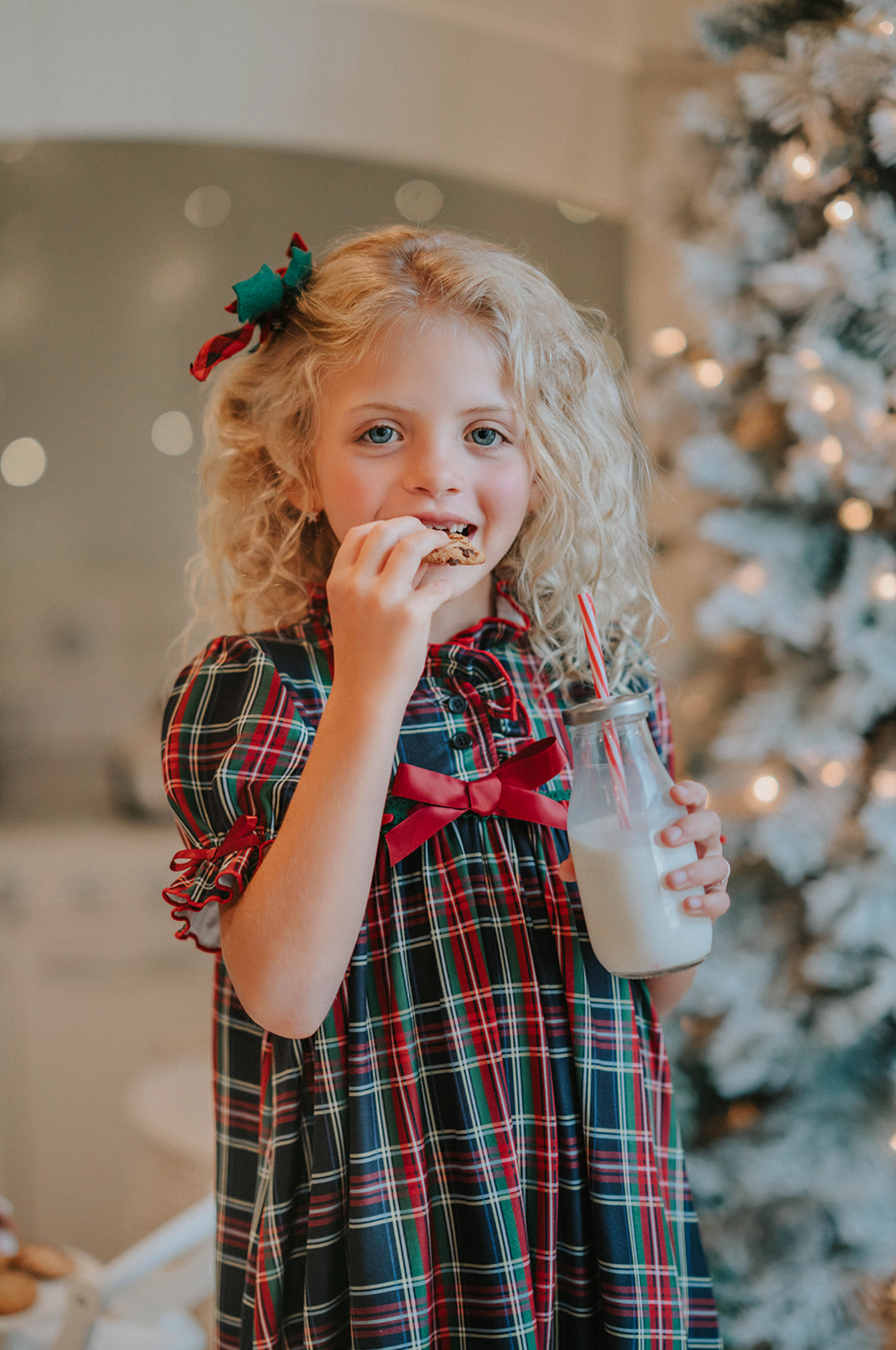 A young girl is wearing The Original Clara Nutcracker Gown in black plaid featuring black plaid ruffles and a red bow. The plaid has white, red and green with a black background. The buttery soft fabric is cozy and sensory friendly. The puff sleeves with ribbon detail and the ruffles at the hem add a traditional classic look.