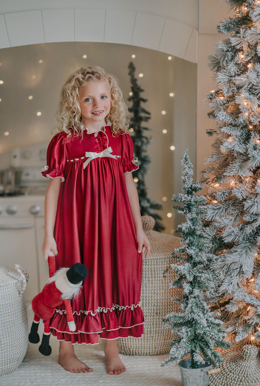 A young girl is wearing The Original Clara Nutcracker Gown in Merry Maroon, featuring maroon ruffles and an ivory bow. The buttery soft fabric is cozy and sensory friendly. The puff sleeves with ribbon detail and the ruffles at the hem add a traditional classic look.