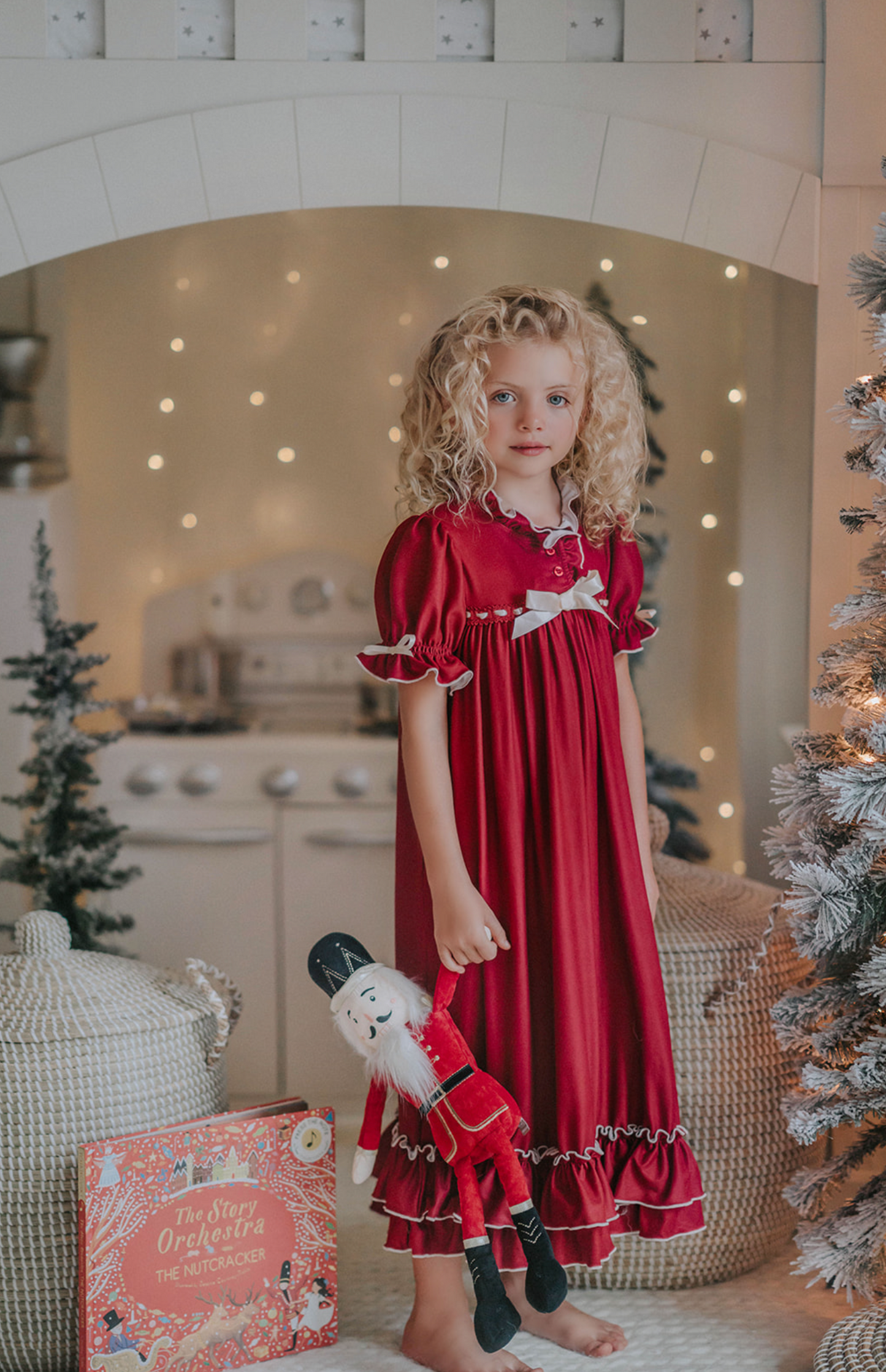 A young girl is wearing The Original Clara Nutcracker Gown in Merry Maroon, featuring maroon ruffles and an ivory bow. The buttery soft fabric is cozy and sensory friendly. The puff sleeves with ribbon detail and the ruffles at the hem add a traditional classic look.