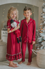 A young boy in buttery soft Unisex Pj's in Merry Maroon holds a nutcracker doll. The traditional 2 piece pajama set has ivory piping and maroon buttons down the front. A girl in matching pajamas with curly hair is partially visible in the background.
