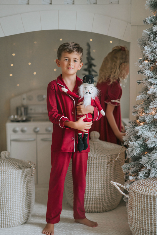 A young boy in buttery soft Unisex Pj's in Merry Maroon holds a nutcracker doll. The traditional 2 piece pajama set has ivory piping and maroon buttons down the front. A girl in matching pajamas with curly hair is partially visible in the background.