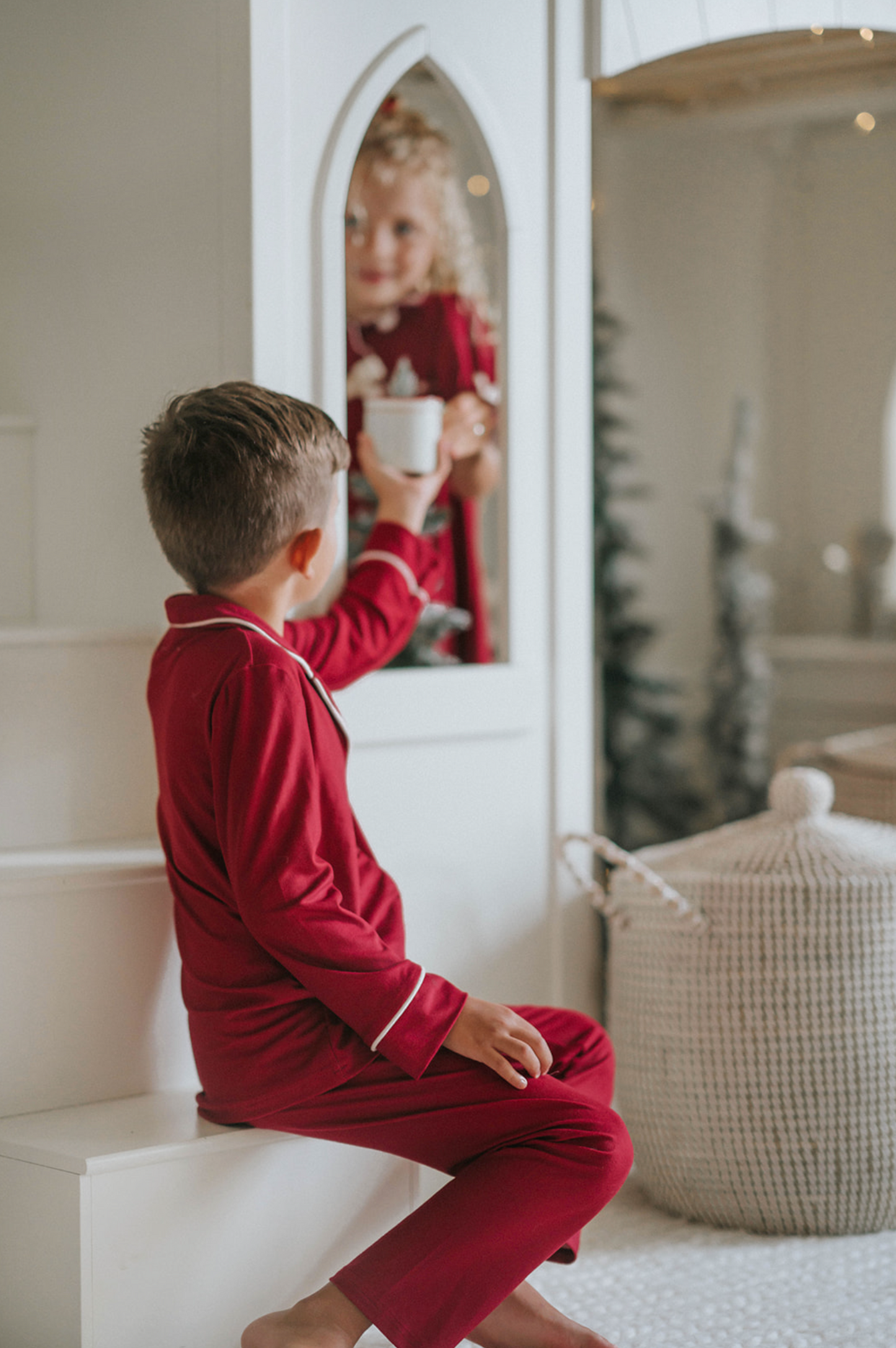 A young boy in buttery soft Unisex Pj's in Merry Maroon holds a nutcracker doll. The traditional 2 piece pajama set has ivory piping and maroon buttons down the front. A girl in matching pajamas with curly hair is partially visible in the background.