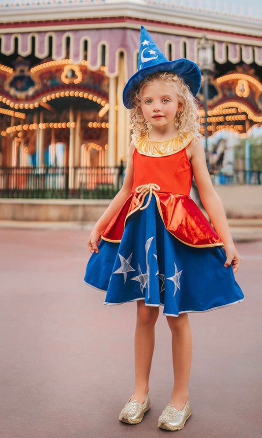 A young girl is wearing our Sorcerer Mouse Twirl Dress and Hat. That wizard hat is blue with silver moon and stars. The dress has a gold collar and red bodice. The skirt has a gold rope bow and a metallic red bustle layer atop a blue twirl skirt. The skirt is adorned with a silver moon and stars.