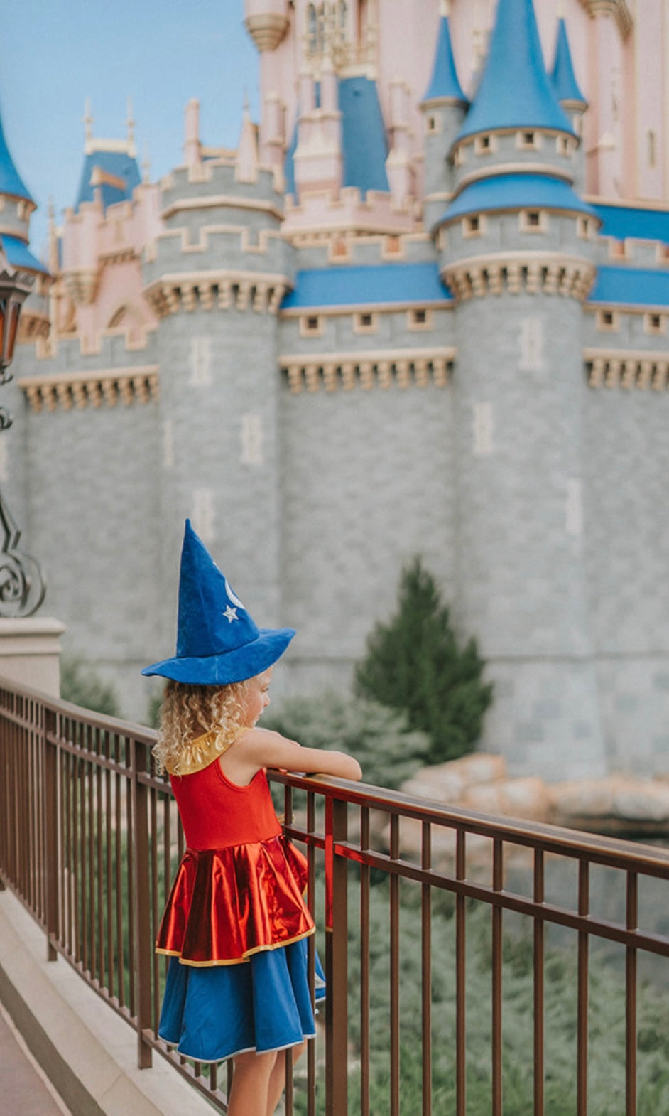 A young girl is wearing our Sorcerer Mouse Twirl Dress and Hat. That wizard hat is blue with silver moon and stars. The dress has a gold collar and red bodice. The skirt has a gold rope bow and a metallic red bustle layer atop a blue twirl skirt. The skirt is adorned with a silver moon and stars.
