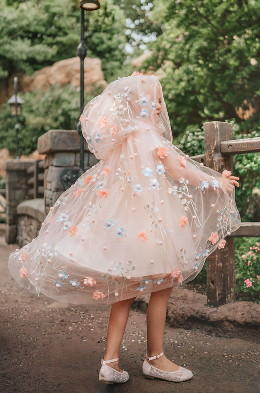 Young girl wearing a very soft pink tulle cape with hood. There are beautiful floral flowers with pink petals and light blue flowers with pink fuzzy centers. Greenery in light green is scattered around the flowers.