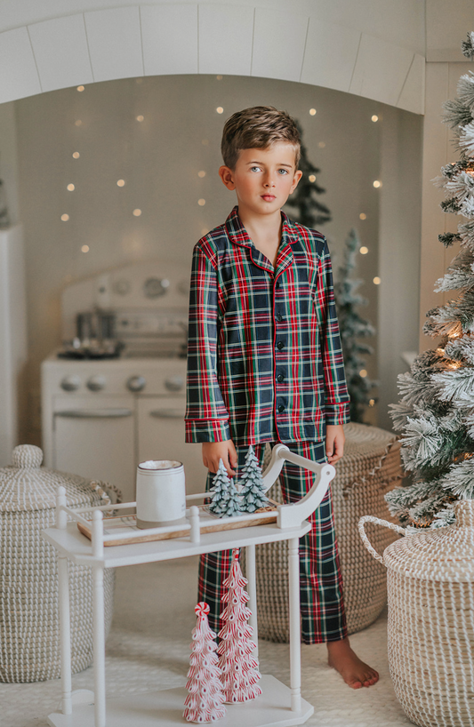 A young boy in Unisex Pj's in Black Plaid stands near a Christmas tree. The classic 2 piece pajama set is a traditional Christmas plaid with black background and red, green and white accents. The top has red piping trim.