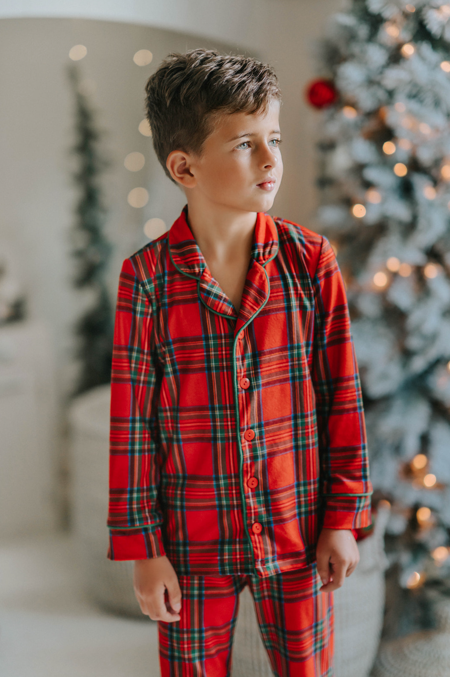 A young child in Unisex Pj's in Red Plaid decorates a Christmas tree. The plaid has green, white and black over a bright red. The classic 2 piece pajamas have green piping and red buttons down the front of the shirt.