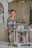 A young boy stands in a festively decorated room wearing Unisex Pj's in White Plaid. The classic 2 piece pajama set is buttery soft fabric. The white background is accented by green, red and black plaid. There is red piping trim on the top.