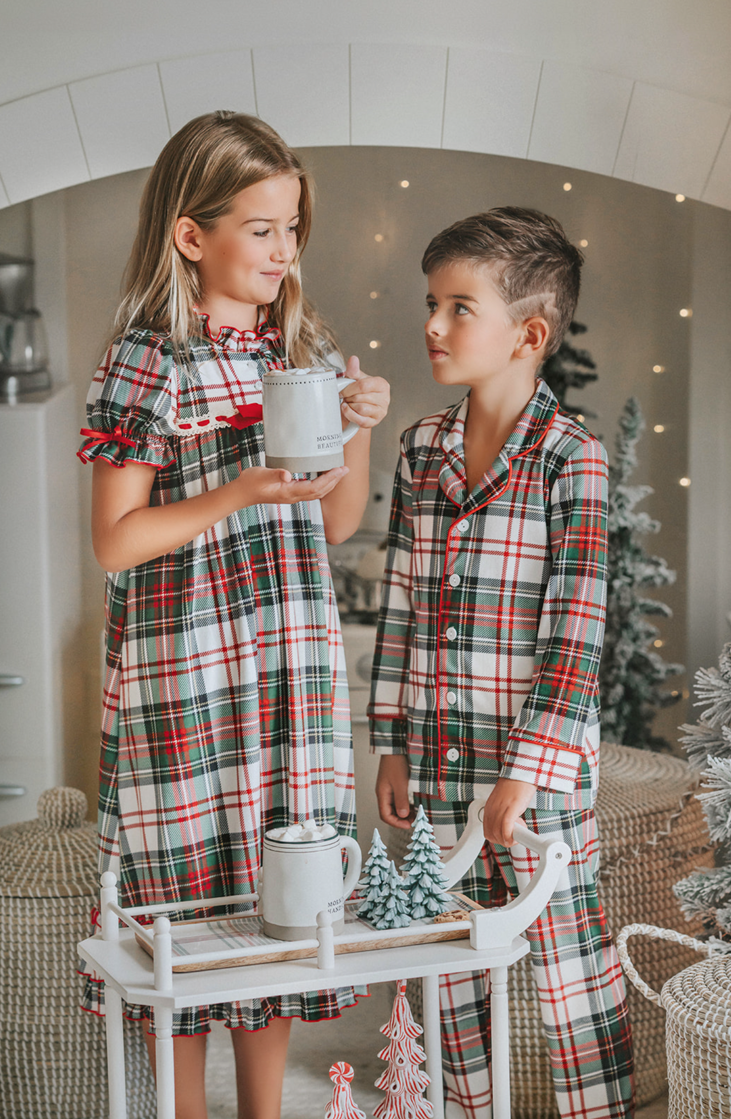 A young boy stands in a festively decorated room wearing Unisex Pj's in White Plaid. The classic 2 piece pajama set is buttery soft fabric. The white background is accented by green, red and black plaid. There is red piping trim on the top.